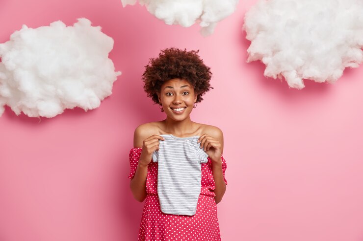 Foto grátis mulher grávida alegre antecipa o bebê, segura o macacão sobre a barriga, quer ter um filho, sorri com alegria, está de bom humor durante a gravidez, isolada em uma parede rosa com nuvens brancas
