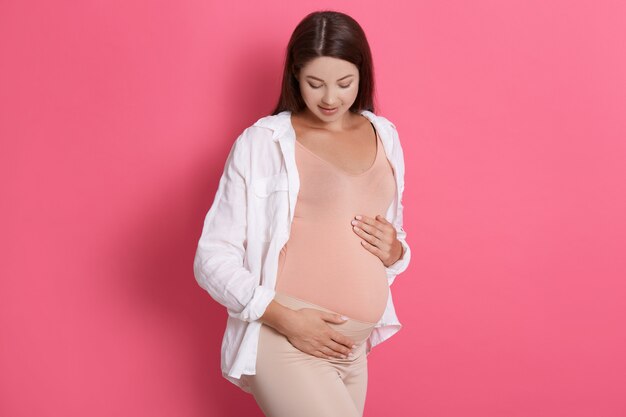 Mulher grávida atraente com cabelo escuro, vestindo leggins e camisa, garota olhando para a barriga dela com muito amor, posando isolado sobre o espaço rosa.