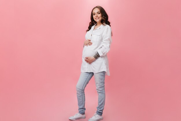 Mulher grávida atraente em camisa longa branca sorri amplamente. Menina morena feliz em poses de jeans em fundo rosa.