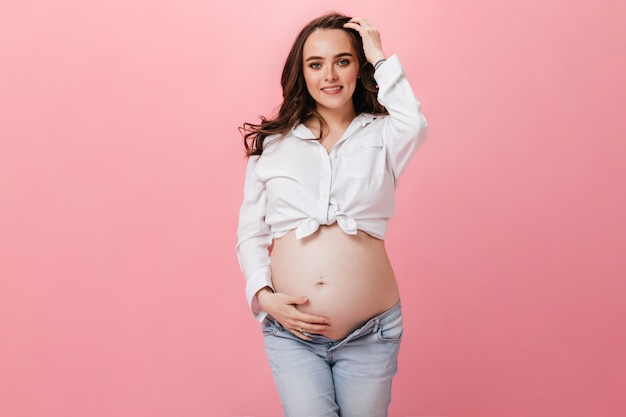 Mulher grávida atraente morena de camisa branca recortada e calça jeans toca a barriga e sorri no fundo rosa isolado.