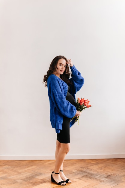 Mulher grávida encantadora no casaco de lã azul contém buquê de tulipas. Jovem feliz em poses de vestido preto com flores isoladas.
