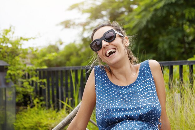 Mulher grávida feliz usando óculos escuros e vestido azul relaxante ao ar livre contra a natureza verde