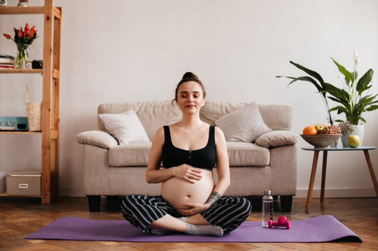 Foto grátis mulher grávida morena alegre gosta de música em fones de ouvido sem fio brancos e toca suavemente a barriga. alegre menina bronzeada em calças pretas e top meditando no tapete roxo.