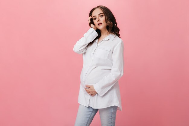 Mulher grávida morena em camisa branca olha para a câmera. Jovem alegre em poses de jeans em fundo rosa isolado.