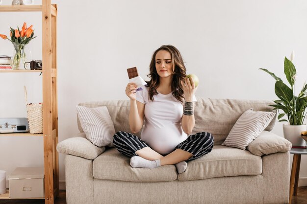 Mulher grávida morena insatisfeita olha para chocolate doce de leite e segura maçã verde enorme e saudável Retrato de menina encantadora descansando no sofá