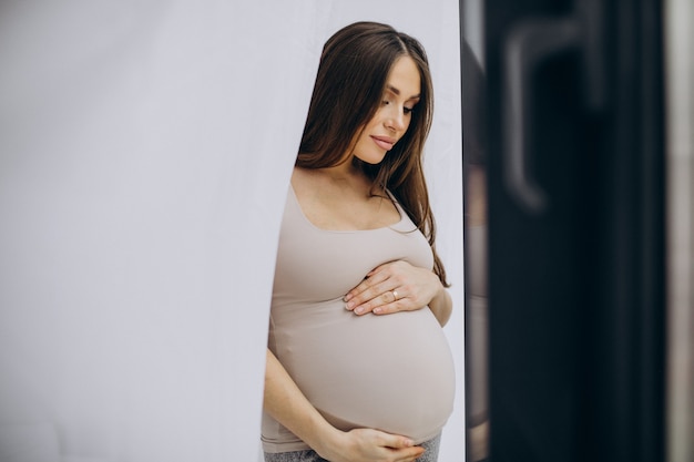 Mulher grávida segurando a barriga e parada perto da janela