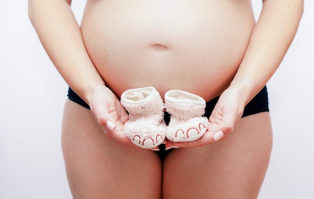 Mulher grávida segurando sapatos de bebê cor-de-rosa na frente da barriga nua