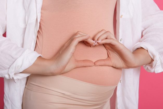 Mulher grávida sem rosto, vestindo roupa bege, posando com o símbolo do coração contra a barriga