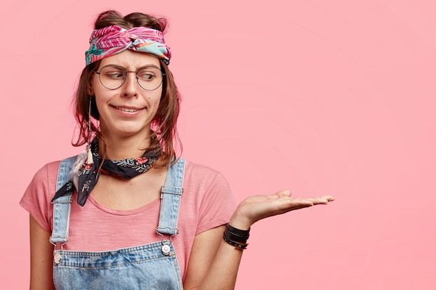 Foto grátis mulher hippie fofa com expressão facial suspeita mantém a palma da mão levantada, demonstra algo contra a parede rosa, vestida com macacão jeans casual, tiara estilosa, finge estar segurando coisa