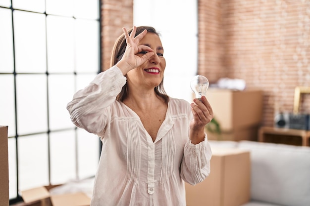 Foto grátis mulher hispânica de meia-idade segurando a lâmpada para inspiração e ideia sorrindo feliz fazendo sinal de ok com a mão no olho olhando por entre os dedos