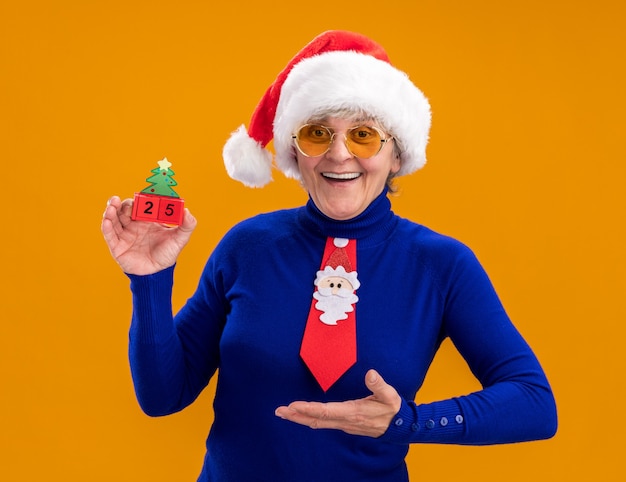 Mulher idosa alegre usando óculos de sol com chapéu de Papai Noel e gravata de Papai Noel segurando e apontando para o enfeite de árvore de Natal isolado em um fundo laranja com espaço de cópia