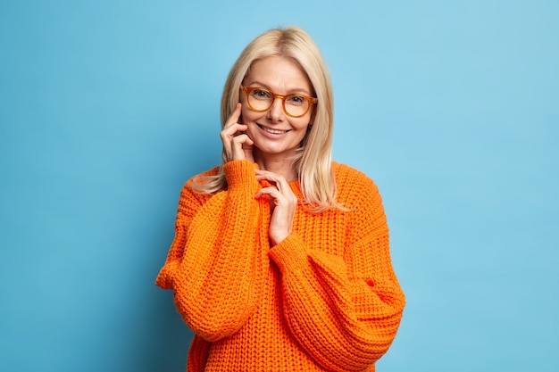 Mulher idosa bonita com cabelo loiro, pele saudável tocando rosto, sorrindo com ternura, usa modelo de suéter de malha laranja