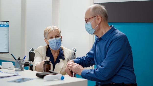 Mulher idosa com deficiência crônica e seu marido na consulta, participando de visita de check-up com especialista médico. Paciente usuário de cadeira de rodas fazendo consulta durante a pandemia de covid 19.