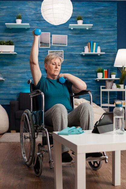 Mulher idosa com deficiência em cadeira de rodas levantando o braço, treinando a resistência dos músculos com halteres