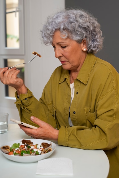 Foto grátis mulher idosa comendo prato de figos em casa e usando smartphone