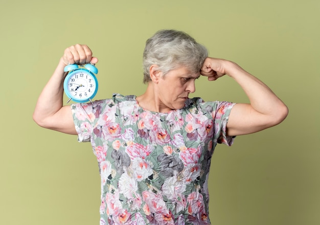 Mulher idosa confiante tensiona os bíceps e segura o despertador isolado na parede verde oliva