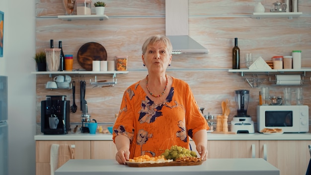 Mulher idosa em uma videochamada na cozinha enquanto a família prepara o jantar. Pov online internet conferência moderna, chat, comunicação, chat, conversa, chamada via webc