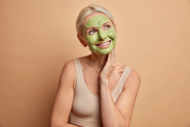 Mulher idosa feliz e satisfeita recebe máscara facial toca pescoço suavemente usa maquiagem mínima e expressão de rosto sonhadora passa por tratamentos de beleza vestida com blusa cortada isolada sobre parede bege