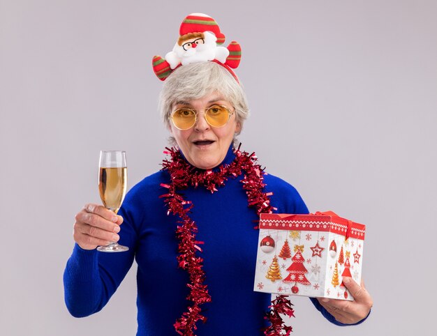 Mulher idosa impressionada com óculos de sol com tiara de Papai Noel e guirlanda no pescoço segurando uma taça de champanhe e uma caixa de presente de Natal isolada na parede branca com espaço de cópia