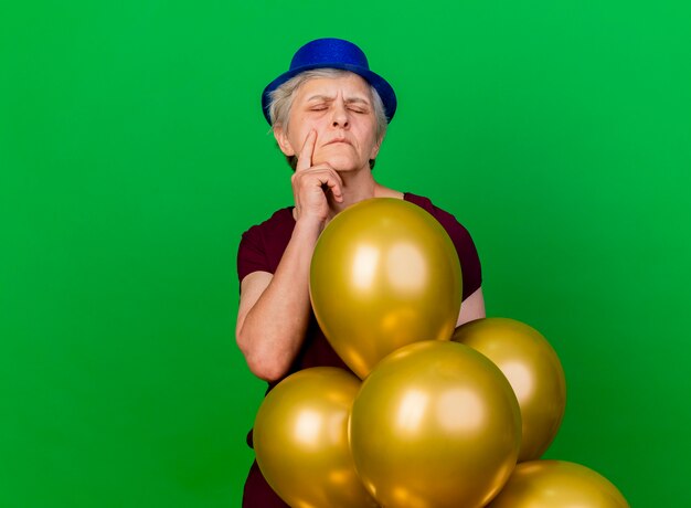 Mulher idosa insatisfeita com chapéu de festa coloca a mão no queixo com balões de hélio isolados na parede verde com espaço de cópia
