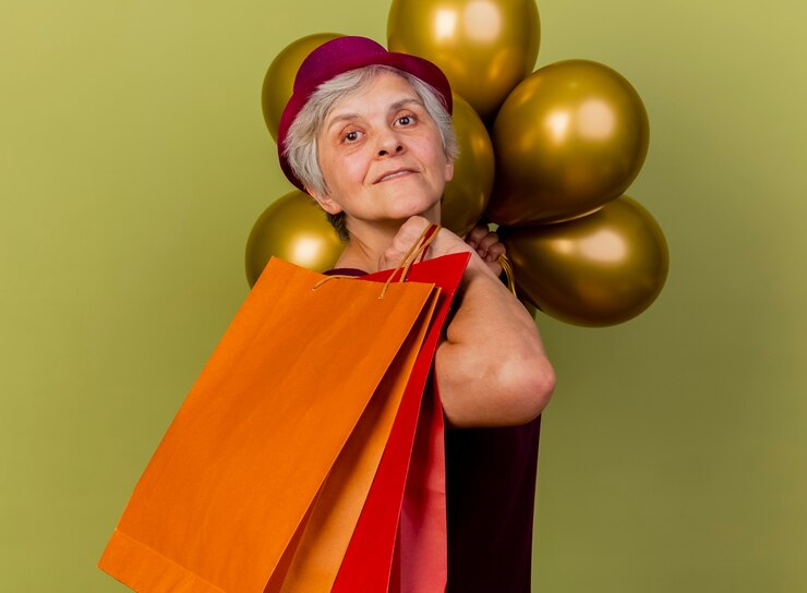 Foto grátis mulher idosa satisfeita com chapéu de festa parada de lado com balões de hélio segurando sacolas de papel isoladas na parede verde oliva com espaço de cópia
