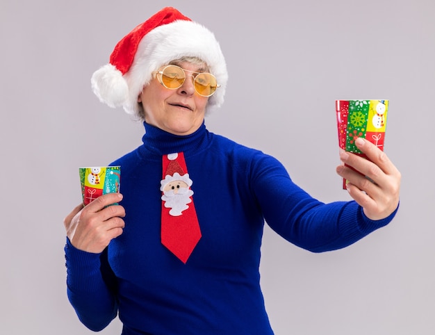 Mulher idosa satisfeita usando óculos de sol com chapéu de Papai Noel e gravata de Papai Noel segurando e olhando para copos de papel isolados na parede branca com espaço de cópia