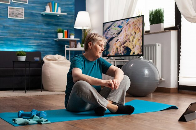 Mulher idosa sentada em posição de lótus no tapete de ioga, treinando os músculos do corpo e reduzindo o peso