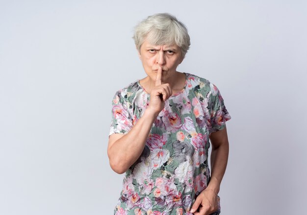 Mulher idosa séria colocando o dedo na boca gesticulando em sinal de silêncio silencioso isolado na parede branca
