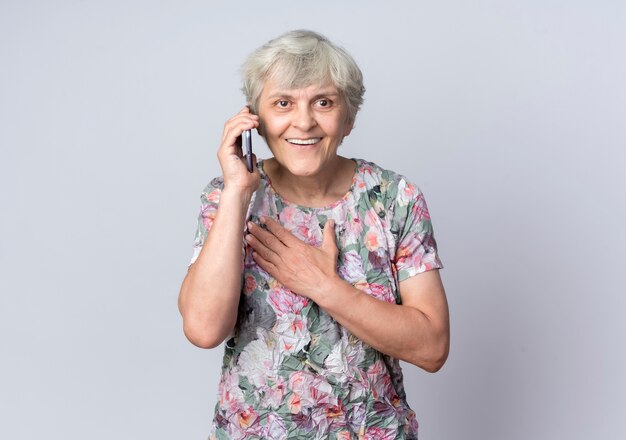 Mulher idosa sorridente coloca a mão no queixo falando no telefone isolado na parede branca