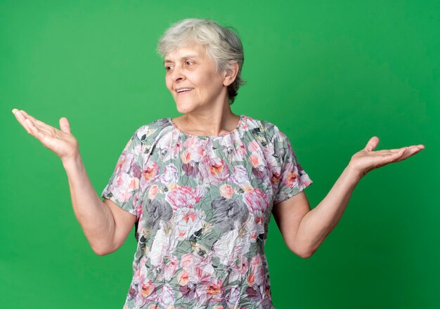 Mulher idosa sorridente em pé com as mãos levantadas, olhando para o lado isolado na parede verde