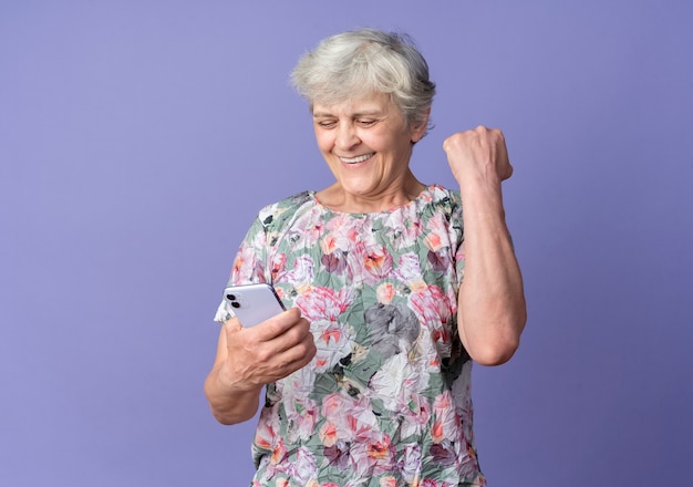 Mulher idosa sorridente levanta o punho olhando para o telefone isolado na parede roxa