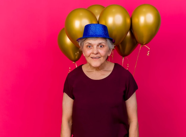 Mulher idosa surpresa com chapéu de festa na frente de balões de hélio olhando para a câmera rosa