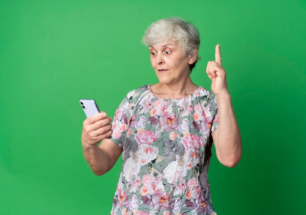 Mulher idosa surpresa olhando para o telefone apontando para cima, isolado na parede verde