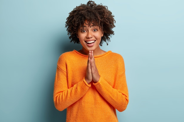 Mulher implorando feliz de pele escura mantém as mãos em gesto de oração, tem olhar suplicante, expressão positiva, pede apoio e ajuda, usa blusão casual laranja, modelos sobre parede azul