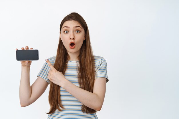 Mulher impressionada apontando para a tela do telefone celular, olhando maravilhada com a câmera, segurando o smartphone horizontalmente, em pé sobre branco