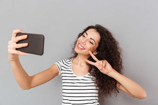 Mulher incrível com aparência caucasiana, fazendo selfie em seu smartphone, sorrindo e fazendo o gesto de vitória com dois dedos