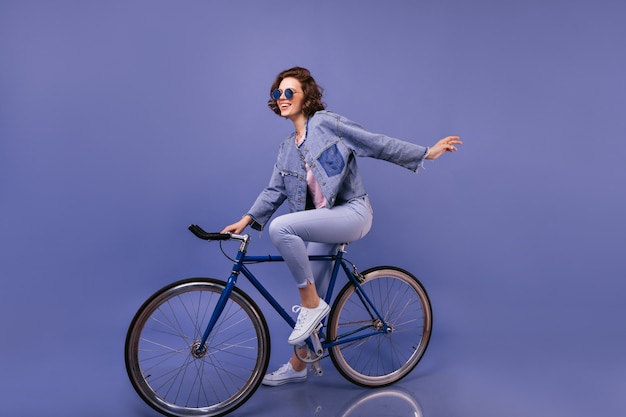 Mulher incrível com roupas de primavera, sentado na bicicleta. retrato interior de uma linda garota de óculos de sol brincando.