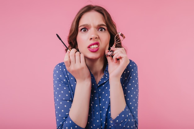 Mulher infeliz com cabelos ondulados, fazendo a maquiagem antes do encontro. menina nervosa em um traje azul cachos cílios na parede rosa.