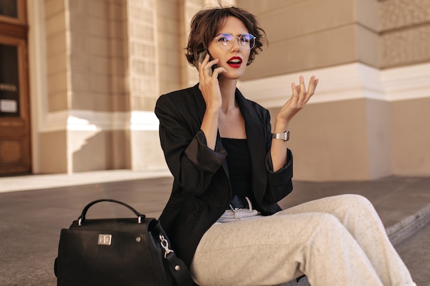Mulher infeliz de jaqueta preta e óculos fala no telefone do lado de fora. Mulher moderna, com lábios vermelhos e cabelo encaracolado, senta-se ao ar livre.