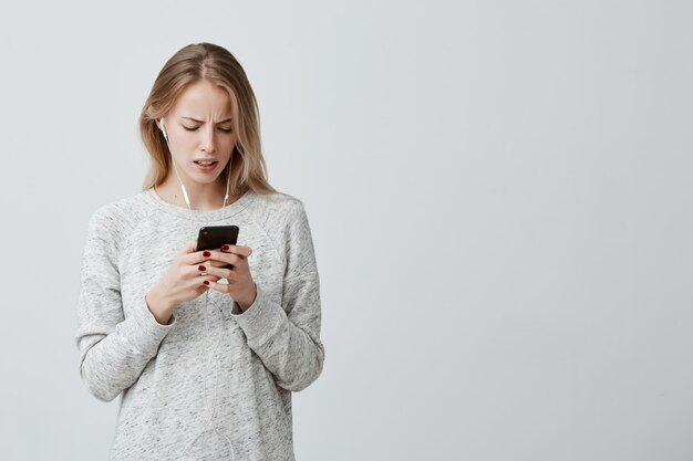 Mulher insatisfeita com cabelos loiros tingidos, vestida casualmente com fones de ouvido brancos, segurando o smartphone recebendo mensagem sendo chocada ao esquecer uma importante reunião com parceiros de negócios