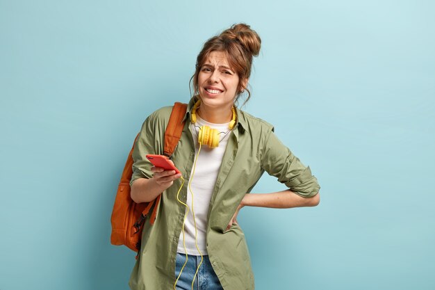 Mulher insatisfeita dá um sorriso afetado, mantém a mão nas costas, sofre de dores terríveis enquanto carrega uma mochila pesada, vestida casualmente