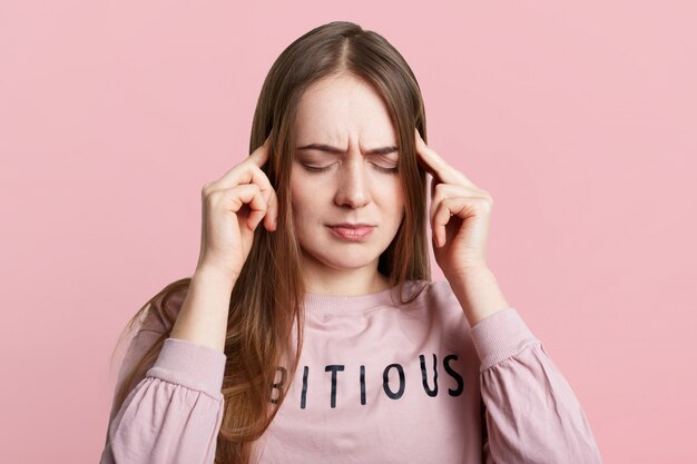 Mulher intrigada mantém os dedos nas têmporas, mantém os olhos fechados, vestido casualmente, tem dor de cabeça após o trabalho mental a noite toda