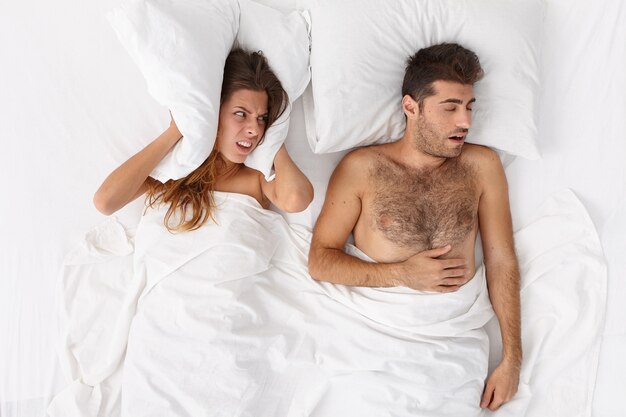 Mulher irritada tapa ouvidos, cobre travesseiro, olha com raiva para marido que ronca, não consegue dormir, fica irritada, tem problemas para dormir, deita em cama branca. Homem tem problemas de apnéia do sono