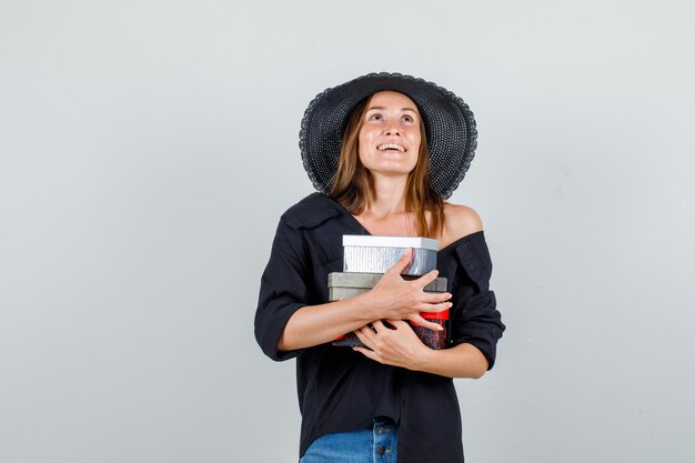 Mulher jovem abraçando caixas de presente enquanto olha para cima com camiseta, shorts, chapéu e parece alegre