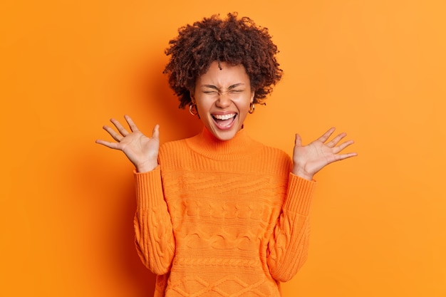 Mulher jovem alegre e positiva levanta as palmas das mãos sente-se muito contente e expressa alegria vestida com um macacão casual isolado sobre a parede laranja