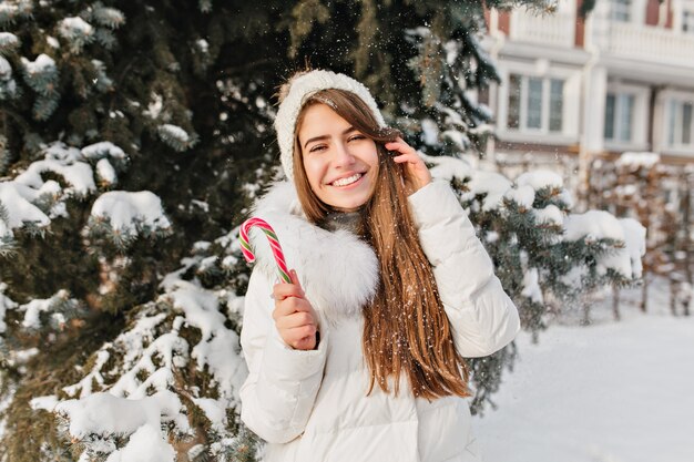 Mulher jovem alegre engraçada com pirulito na cidade de inverno. Bom humor, roupas quentes, neve caindo, emoções brilhantes, expressões, ano novo e época de natal