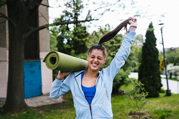 Mulher jovem alegre esportes caminhando no parque urbano, segurando o tapete de fitness.