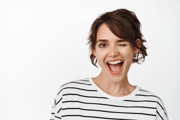 Mulher jovem alegre piscando para você, sorrindo confiante, pele limpa e brilhante de uma menina linda morena, em pé com uma camiseta branca