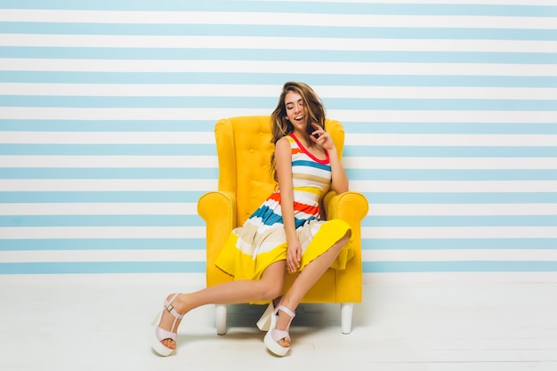 Mulher jovem animada com lindo sorriso relaxando em uma poltrona confortável. linda garota de cabelos compridos, usando um vestido da moda e sapatos de salto, sentada na mobília amarela com os olhos fechados.