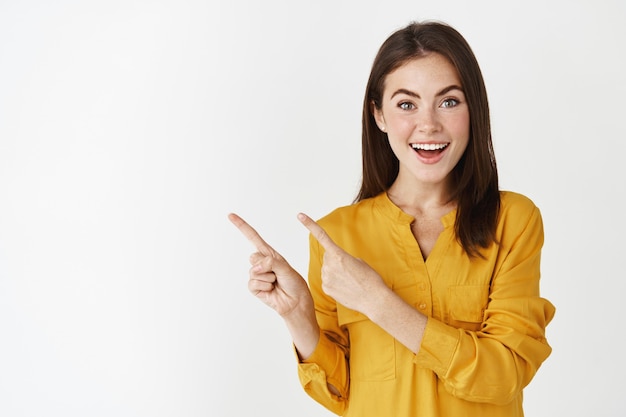 Mulher jovem animada mostrando o banner, apontando o dedo para a esquerda e sorrindo para a câmera, admirada com a parede branca